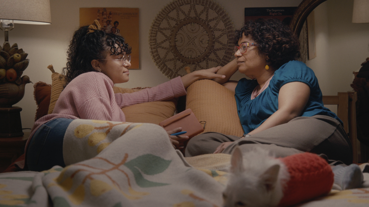 A Black woman with long wavy hair, wearing a pink sweather and jeans, sits on a couch facing her mother, a Black woman with wavy hair, wearing a hunter green shirt and brown pants. From Aurora Brachman’s ‘Still Waters.’ Courtesy of DOC NYC