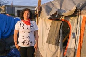 A homeless woman wearing a RIP 6ixx shirt stands next to an encampment