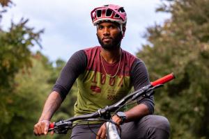 A Black man wearing a helmet sits on a bike. There are trees in the background.