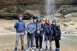 Five donor-conceived siblings wearing winter clothes stand in front of a waterfall. 