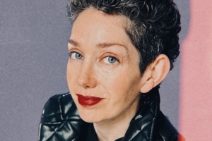A woman with white skin smiles at the camera. She has short, curly salt-and-pepper hair, blue eyes, red lipstick, and is wearing a quilted black leather blazer. 