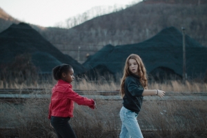 Two young girls playing together against a backdrop of mountains, weeds, and mounds of dirt.