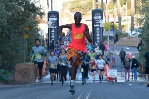 A man wearing sunglasses, shorts, and a neon colored top sprints ahead of a group of runners on a paved street