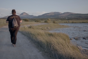 A person wearing all black with a dark gray backpack walking down paved road with a grassy water area to their side and mountains off in the distance.