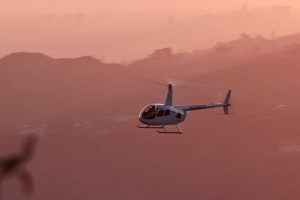 A helicopter is flying past the hazy silhouettes of the Hollywood Hills in a pink sunset.