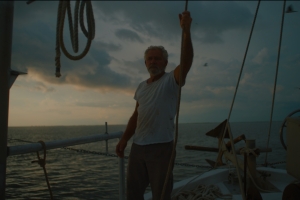 A nearly silhouetted silver-haired man stands on the bow of a boat, holding onto a rope above his head. Behind him is a cloudy dusk sky and an expanse of water.