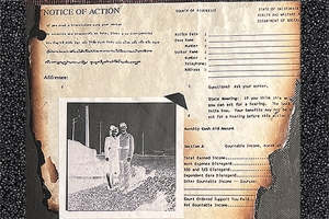 A collage-style painting. It is a black painted canvas with a document affixed on the canvas that reads Action. The document has burned edges. On top of the burned document there is a 1940s photograph mounted with a photo corner. The photograph is printed as a negative and has the ghost-like images of a woman and man posing together in their dress military uniforms.