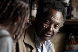 An older Black man wearing glasses is interviewing a blurry figure in the foreground