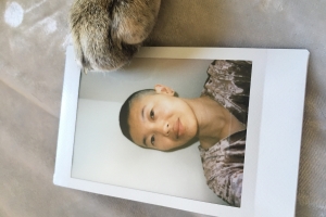 A photo of an androgynous person of Asian descent with a light smile at the camera. They have a dark shaved head and brown eyes and are wearing a pink velvet v-neck. The photo frames a photo print from a Polaroid camera; the print is lying flat on a silver texture, with a cat's paw gently lying on the top right corner of the print.