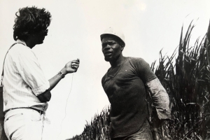 black and white photo from the Victor Jara Collective featuring a man holding a small microphone up to another man wearing a hard hat standing against a row of brush