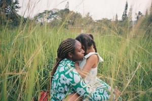 Two black girls, one older and one younger, hug in field of grass.