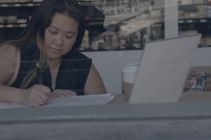 A woman with long hair and a dark top seen through a window writing in front of a laptop