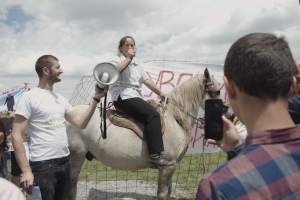 Person on a horse talking to a crowd with a banner in the background. With a person handing them a megaphone