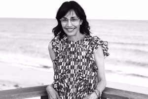 In black and white, Andrea Nevins stands on a balcony with the ocean behind her, smiling brightly.. 