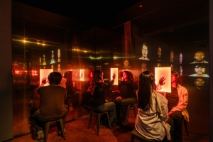 Visitors, framed in an orange glow, put their hands up to a wall-panel as part of the "In Search of Us" exhibit.
