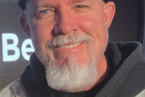 middle aged, caucasian man smiling with a gray goatee and blue eyes wearing a black baseball cap and a black hoodie with a gray sport coat