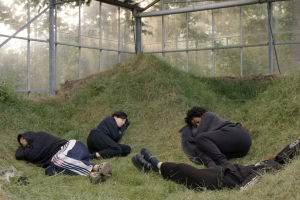 Four people are pictured, curled up in grass inside what appears to be a greenhouse. 