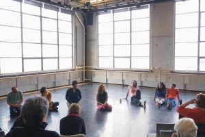 A group of people sit in a circle in a large dance studio. 
