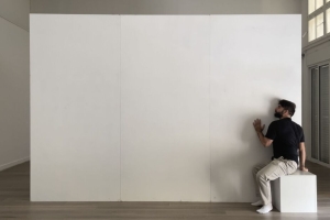 Alaa Minawi sits on a white box, with his ear pressed against a white wall in the center of a bare room.