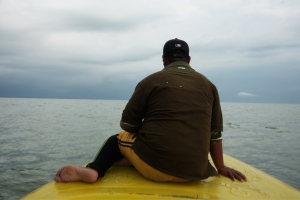 Barefoot and steady, a man sits on the bow of a yellow boat, his back to the camera, shirt weathered by time. He faces the horizon as the vessel cuts quietly through the water. The lake stretches endlessly under heavy clouds, evoking both solitude and endurance.