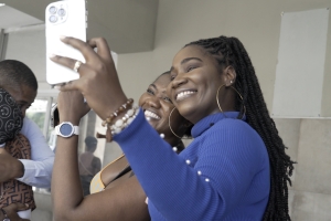 Vanessa and Tamara stand close together, smiling brightly as they take a selfie on a smartphone. Vanessa, in a royal blue top with long braids and hoop earrings, holds the phone out in front of them, while Tamara, dressed in a patterned outfit, leans in beside her. Both women radiate joy, their wide smiles filling the frame. Just behind them, Vanessa’s husband embraces someone tightly, his face pressed against a shoulder. The moment is tender yet bittersweet—Vanessa and Tamara are capturing one last photo t