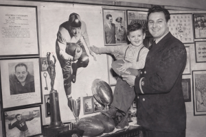 Black and white photo of Sid holding his son Bobby. They are in front of a poster of Bobby as a quarterback.