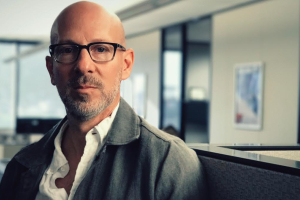 A man with white skin looks at the camera. He wears glasses, a grey jacket, white shirt and is bald and has a light beard. Headshot of Joshua Seftel