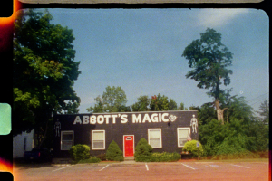 a 16mm film still of a low, black painted brick building with skeletons painted on the front labeled "Abbot's Magic" in large white lettering
