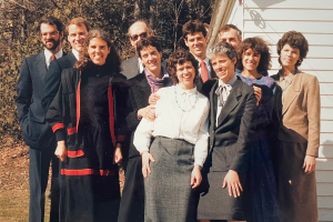 A group of ten siblings stand outside against a white house in late 1980s formal attire. They smile and squint in the sun. Their father, elderly, stands in the back and is mostly obscured.
