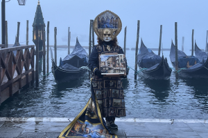 A masked figure in a Carnevale costume stands in front of gondolas and a dock at the edge of the island of Venice, Italy