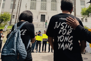 A peaceful outdoor rally taking place on the steps of Los Angeles City Hall. In the foreground, two people are seen from behind, wearing black t-shirts that read "Justice for Yong Yang." Behind them, a diverse crowd of participants of various ages, some wearing hats and sunglasses, hold signs that read "Mental Health Lives Matter" and "Koreans United for Justice."