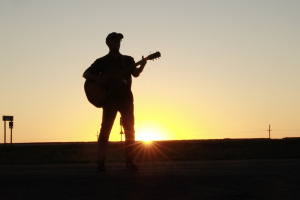 A lone man plays an acoustic guitar on the side of a freeway, silhouetted by the sunset at his back