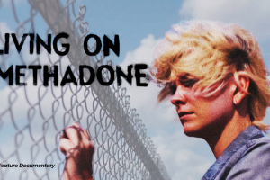 The image shows a young person with short, windblown blond hair standing beside a chain-link fence under a bright blue sky with large white clouds. They are wearing a gray denim-style jacket and appear to be looking through or past the fence with a quiet, reflective expression.  The composition feels cinematic and symbolic. The fence creates a strong visual barrier, suggesting themes of separation, confinement, longing, or being caught between two worlds. The open sky behind them adds contrast, giving the i