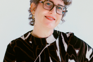 A woman with fair skin smiles at the camera. She has short, curly hair, green eyes, and is wearing a patterned shirt. 