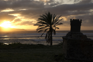 A dramatic sunset over the Molokai'i sea cliffs and ocean waves. In the foreground, a silhouetted stone tomb rests near the shore with a solitary palm tree  leaning slightly in the wind.