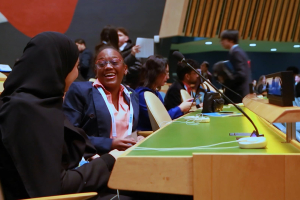 A candid photograph inside the United Nations General Assembly Hall captures two young delegates sharing a joyful moment between sessions. One student, wearing a black hijab, sits facing another delegate in a suit and conference lanyard, who is laughing openly. Around them, other students talk, move between seats, and prepare for proceedings, with microphones and the iconic General Assembly desks visible throughout the frame.  The image captures the heart of The Delegates: young people participating in seri
