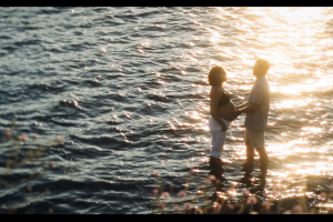 A beautiful, serene scene of a man and woman standing in the water at sunset as the man holds the woman's pregnant belly.