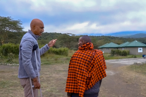 A tall man standing on crutches talks to a Maasai elder wearing a traditional shuka with small buildings behind them.