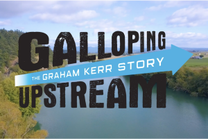 A cinematic aerial view of a winding river surrounded by lush green forests and open farmland under a blue, cloud-filled sky. Bold, distressed black lettering reads “GALLOPING UPSTREAM,” crossed by a bright turquoise arrow containing the white subtitle “THE GRAHAM KERR STORY.” The design feels adventurous, energetic, and inspirational, suggesting a journey of perseverance and discovery.