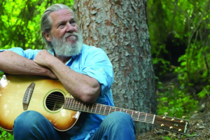 This image contains actor Jeff Bridges with a guitar, sitting in a forest looking up at the trees.