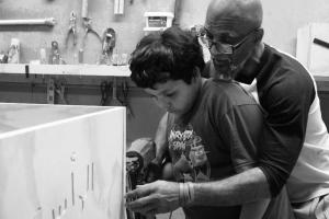 :A  bald Black man with glasses teaches a young boy with black hair how to engrave a casket with a drill.