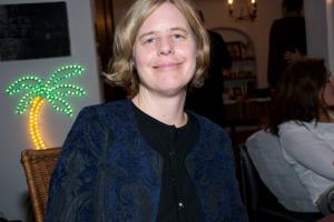 The late Beth Bird sits in a restaurant, smiling; she has short blond hair, and is wearing a blue jacket over a black shirt.