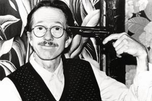 A man wearing glasses with a closed-lip smiles holds a gun up to his temple. 