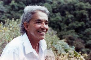 Chavela Vargas is a queer Latin American singer with grey hair. She is wearing a white shirt and is smiling. She is surrounded by trees and shrubs. Image from Catherine Gund and Daresha Kyi’s ‘Chavela.’ Courtesy of Alicia Elena Pérez Duarte, Aubin Pictures.