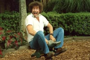 Bob Ross is white artist with a brown afro and a mostly grey beard. He is riding a bouncy alligator ride in a playground. He is wearing a white shirt and blue jeans. Image from Joshua Rofé’s ‘Bob Ross: Happy Accidents, Betrayal & Greed.’ Courtesy of Netflix. 