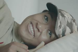Naomi Osaka, a Black and Asian bi-racial woman, lying on her side and smiling. She is wearing a camo hat. A still from Garrett Bradley’s docu series ‘Naomi Osaka’, which was the opening night film at AFI Docs. Courtesy of Netflix.