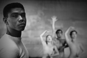 Black-and-white image of a young Alvin Ailey. He is a Black man with short hair wearing white t-shirt. Behind him are three dancers. Image from Jamila Wignot’s “Ailey”. Courtesy of NEON.