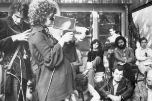 Black-and-white image of French feminist filmmaker Carole Roussoupolos, whose films are now streaming on “Another Screen.” In the photo, she is pregnant and is filming using a handheld camera, with people looking on. Courtesy of Another Screen; Centre Audiovisuel Simone de Beauvoir, Paris.