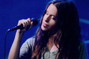 Singer Alanis Morissette, a white woman with long black hair, is performing on stage against a blue lit background; she is holding a microphone, and her eyes are closed, From Alison Kaynman’s ‘Jagged.’ Courtesy of TIFF.