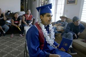Denilson Garibo is a young man in a blue graduation gown. He is wearing a graduation hat and has a garland of white plastic flowers around his neck. He is surrounded by family members. From Peter Nicks' ‘Homeroom.’ Courtesy of Hulu